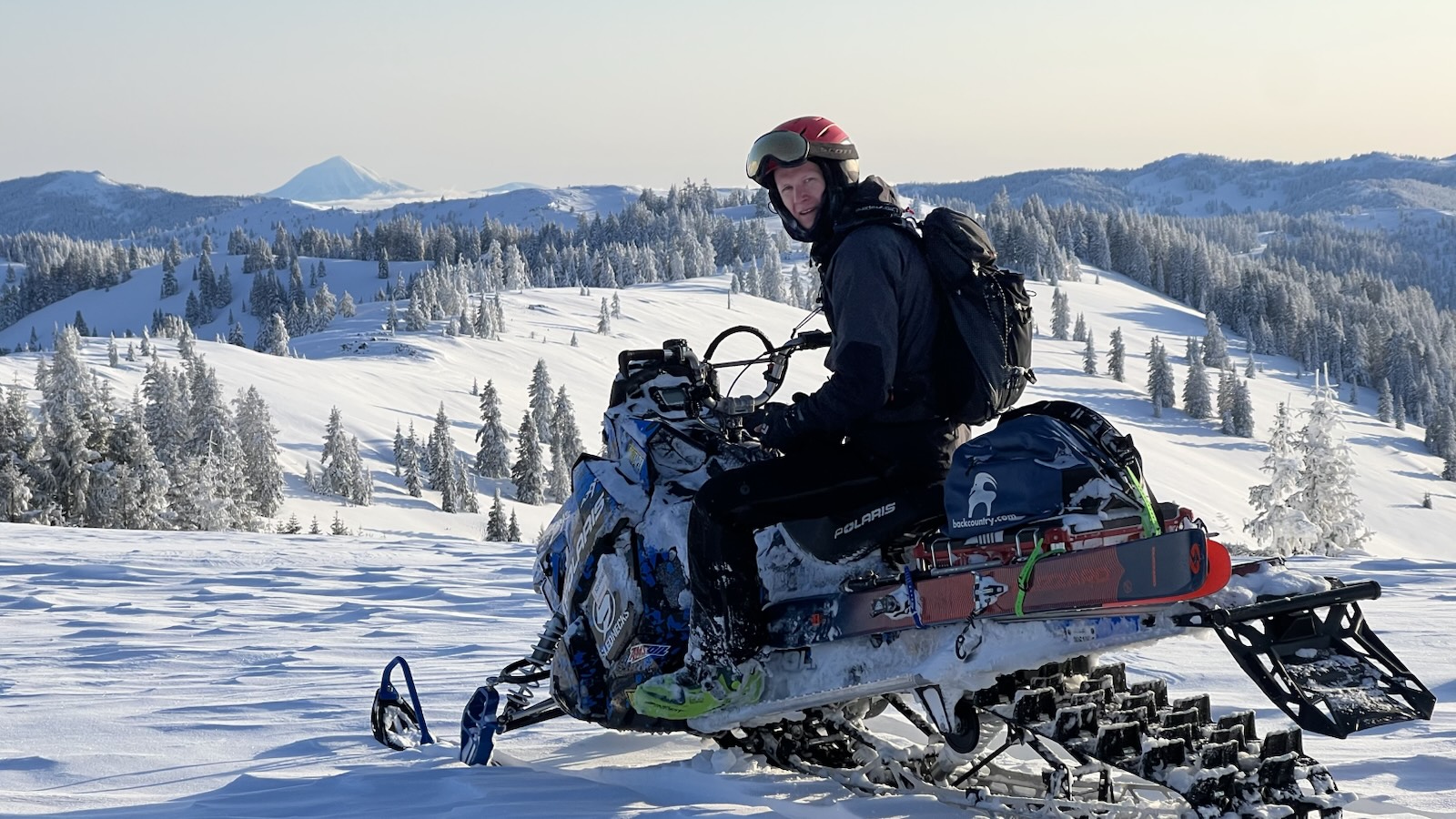 Kevin on a snowmobile loaded with skis, with snowy ridges and Mt. McLoughlin behind